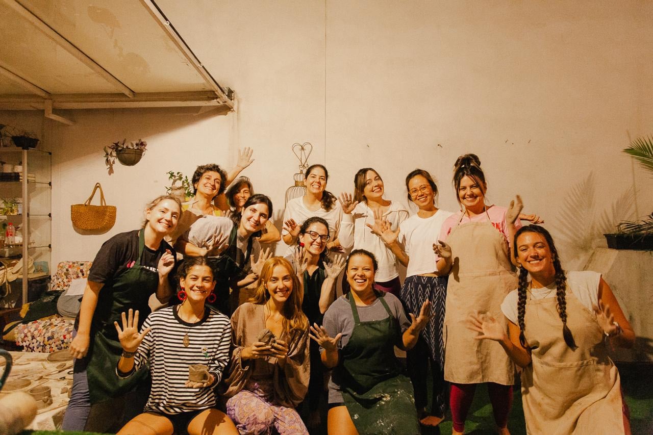 Foto de grupo de uma turma de cerâmica: pessoas de avental erguem as mãos manchadas de barro, sorrindo, em um ateliê de parede salmão.