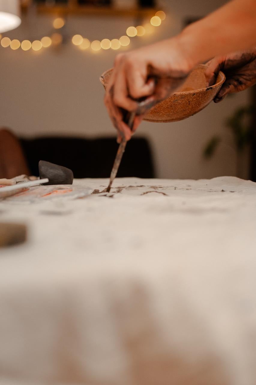 Mão aplicando engobe com um pincel a partir de uma tigela de terracota, com um fio de luzes desfocadas ao fundo.