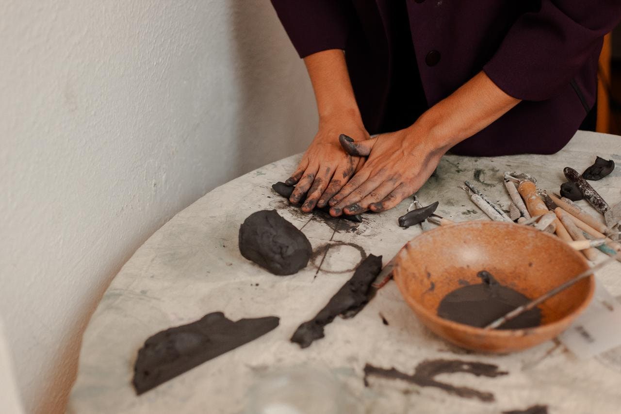 Mãos amassando um bloco de barro escuro sobre uma mesa de mármore branco, ao lado de uma tigela de terracota e ferramentas de madeira.